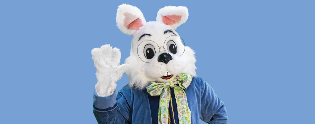 Person in a white bunny costume with glasses and a colorful bow tie waves, set against a solid blue background.