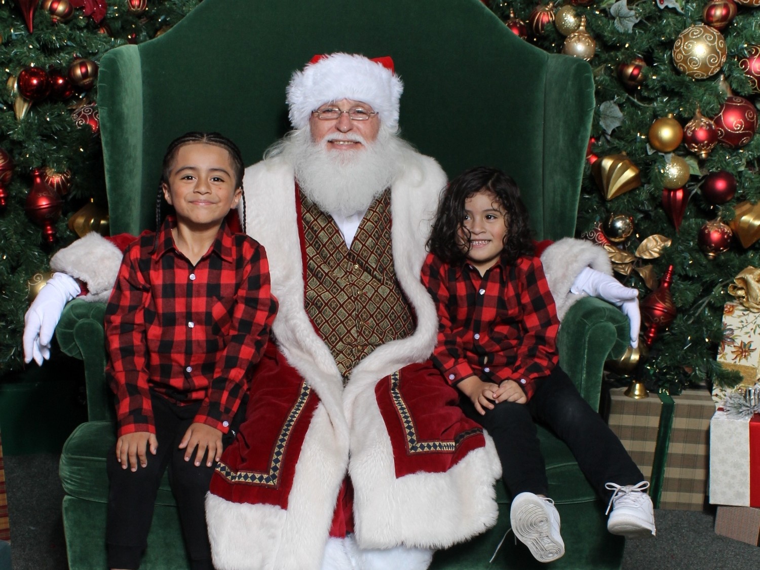 Two children in matching red plaid shirts sit on a green velvet chair next to Santa, surrounded by festive holiday decorations.