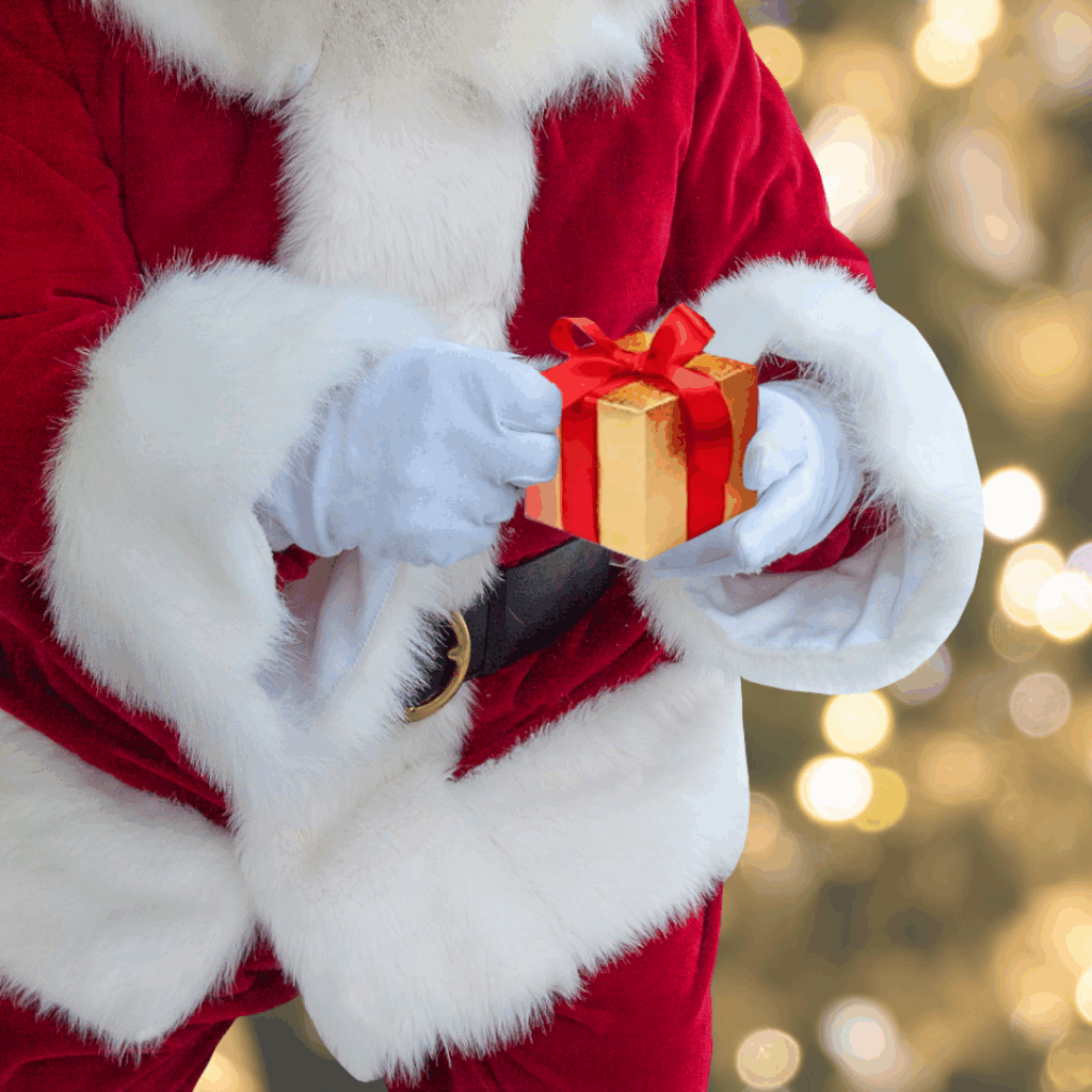 Santa Claus holding a small gold gift box with a red ribbon, against a blurred background of festive lights. The scene conveys warmth and holiday cheer.